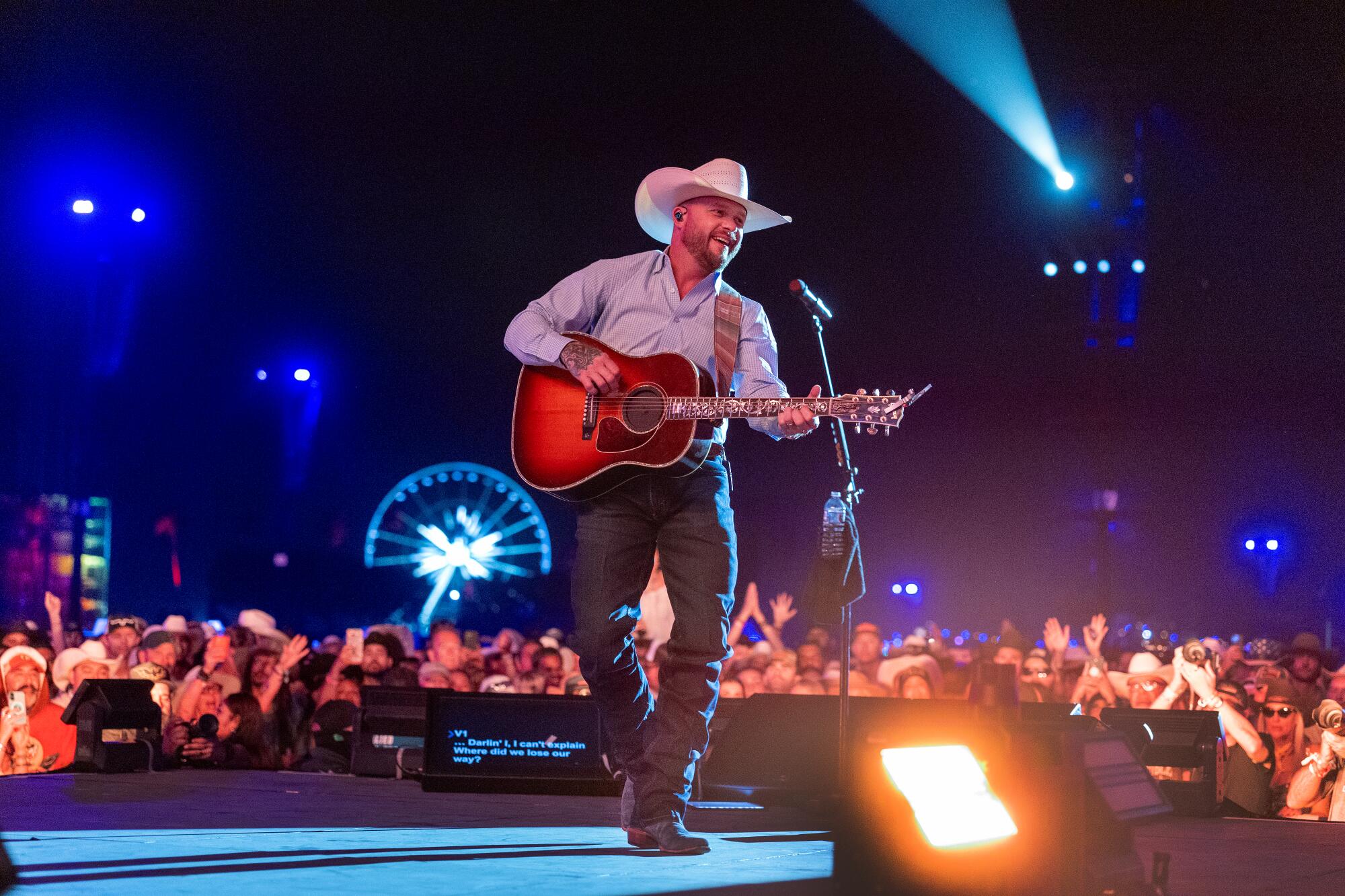 Cody Johnson performs with a guitar at Stagecoach with fans and a lit-up ferris wheel behind him