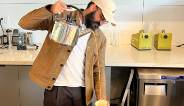A man wearing a beige jacket and cap pours tea from a tall metal kettle into a green paper cup in a modern kitchen.
