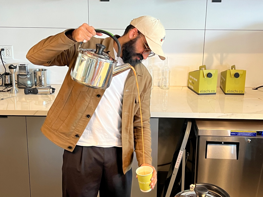 A man wearing a beige jacket and cap pours tea from a tall metal kettle into a green paper cup in a modern kitchen.