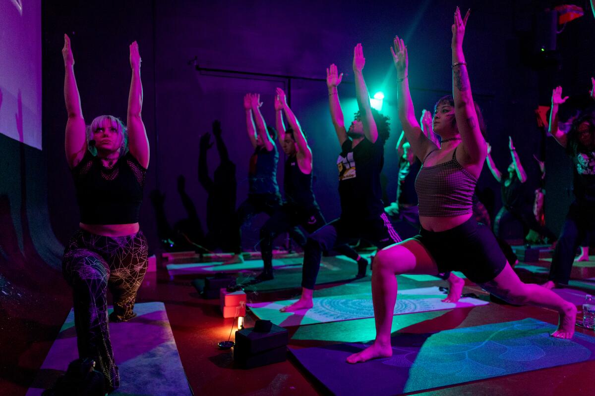 In a dark room with a blue spotlight, people stand on mats in a forward lunge with their arms above their heads.