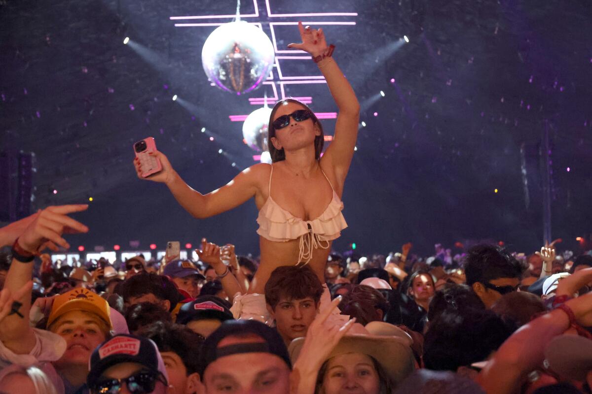 A woman in a bikini top and sunglasses sits on a man's shoulders in a crowd of young people.
