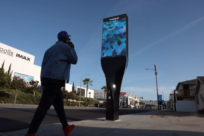 A low angle view of a person walking down a sidewalk towards a vertical digital billboard. There are homes and apartments on the side of the sidewalk and large buildings and a stadium in the other side.