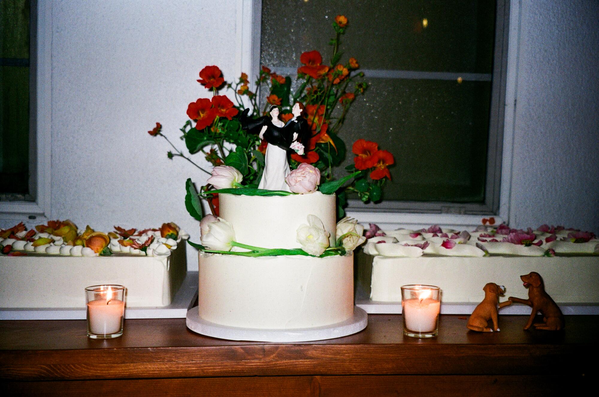 A cake covered in fresh flowers with a wedding topper of a woman carrying a man and a dog figurine at right.