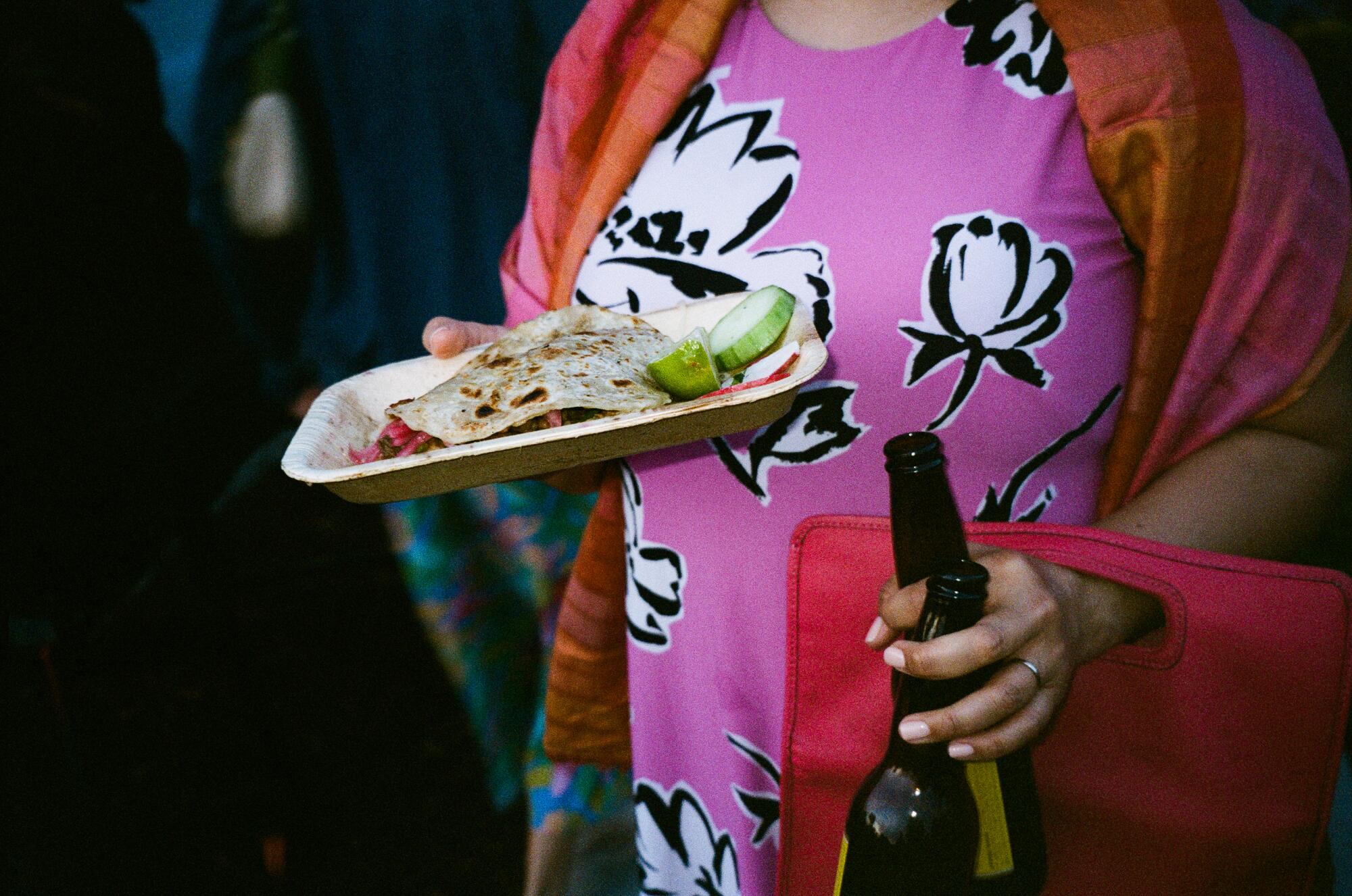 A woman carries a taco on a plate in one hand and two beer bottles in another.