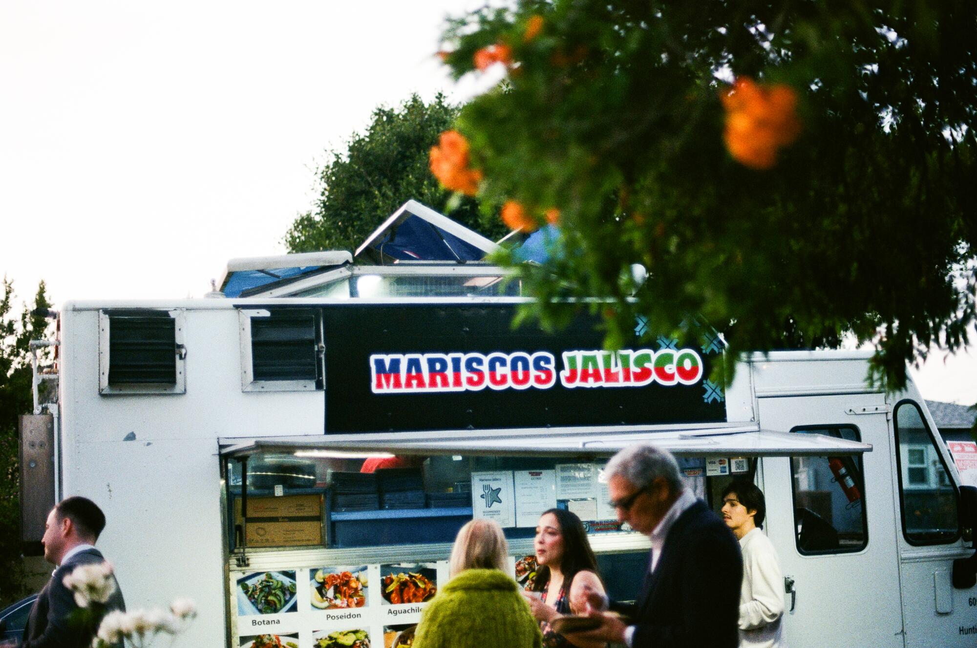 People in wedding attire stand in front of a white food truck.