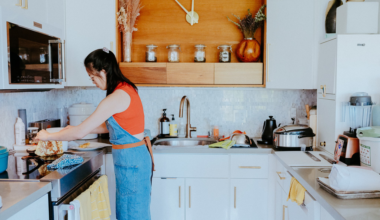 A woman wearing glasses, a red sleeveless top, and a denim apron stands in a kitchen with a metal mixing bowl and fresh produce on the counter.