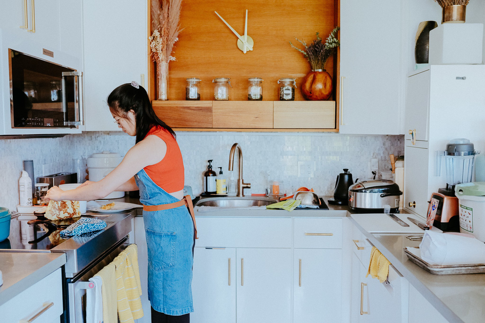 A woman wearing glasses, a red sleeveless top, and a denim apron stands in a kitchen with a metal mixing bowl and fresh produce on the counter.