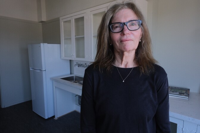 A woman, with light skin tone and black clothing, stands in the kitchen of a Hollywood apartment building.
