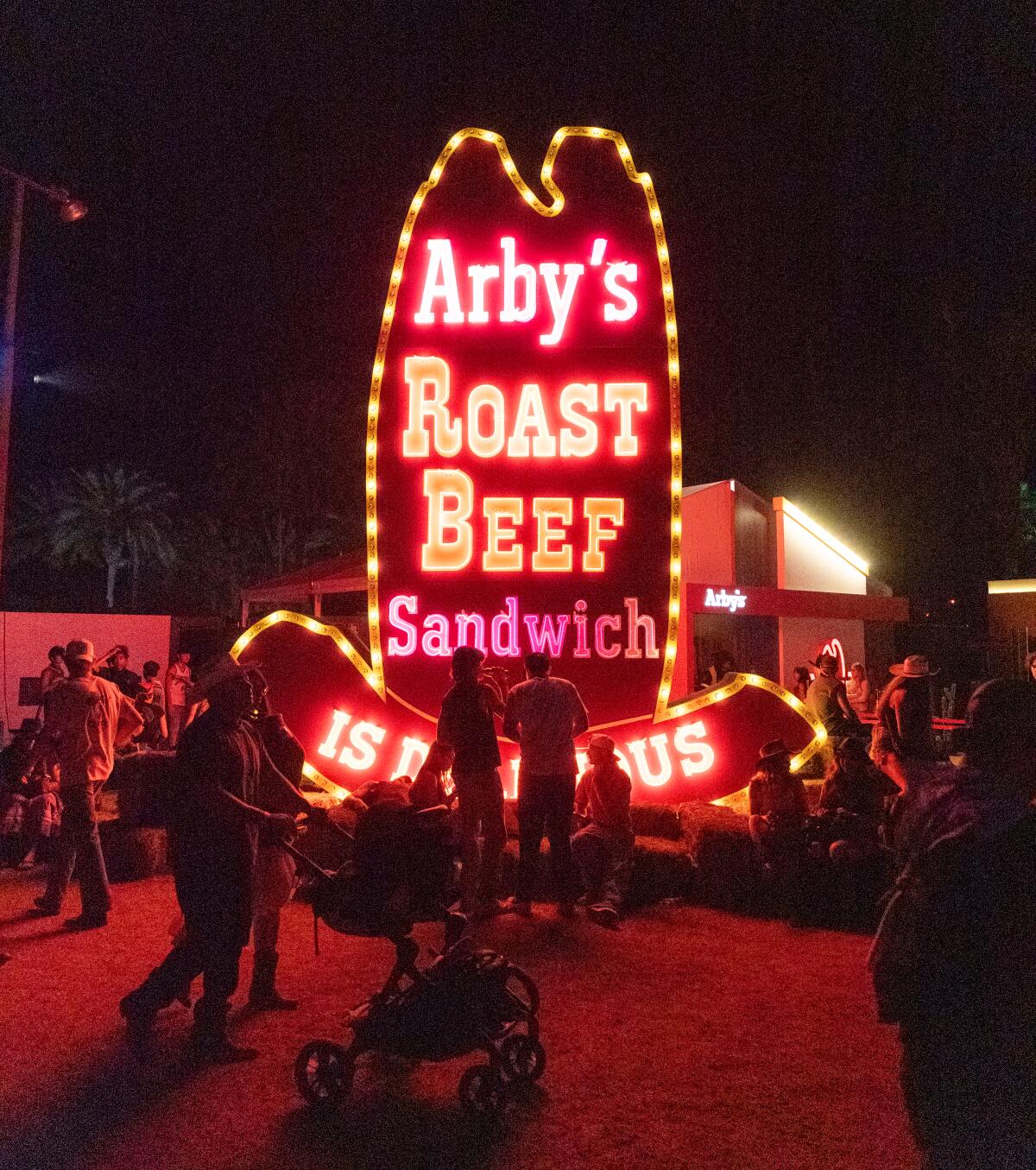 People gather around a lit-up old-fashioned Arby's Roast Beef Sandwich sign shaped like a cowboy hat
