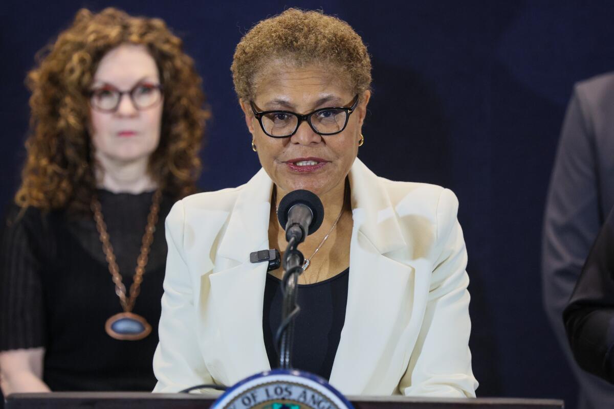 a woman wearing glasses speaks at a mic during a press conference as another woman stands behind her
