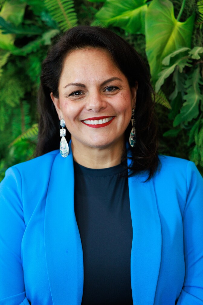 a smiling woman with shoulder-length hair and dangling earrings wearing a light blue blazer over a dark blue blouse