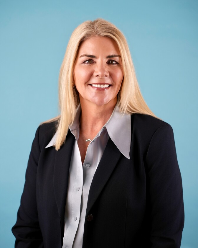 a smiling woman with blond shoulder-length hair wears a dark blazer over a light gray button-up collared shirt