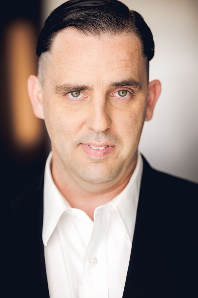 a slightly smiling man with short brown hair and green eyes wearing a dark blazer over a white collared shirt