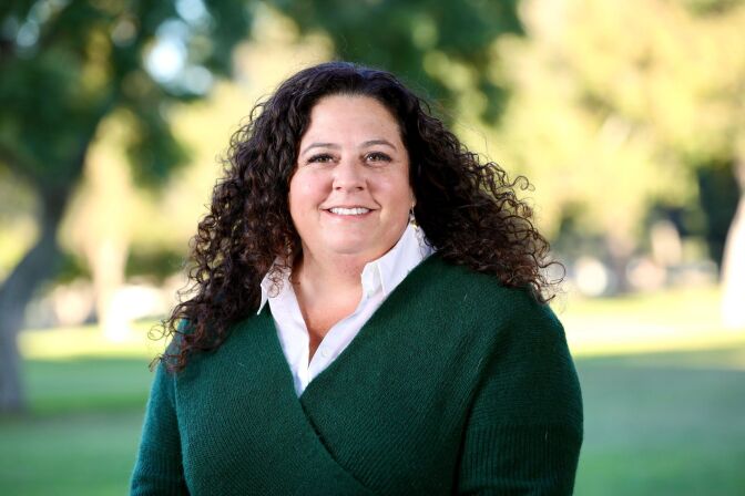 a smiling woman with long curly brown hair wearing a green sweater over a white collared shirt