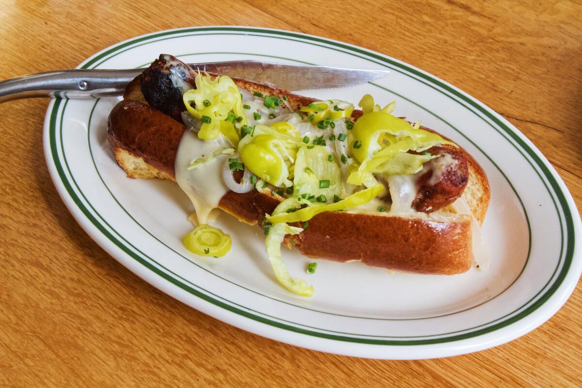 A horizontal photo of a sausage hot dog smothered in cheese and pepperoncini. A French steak knife rests on the plate