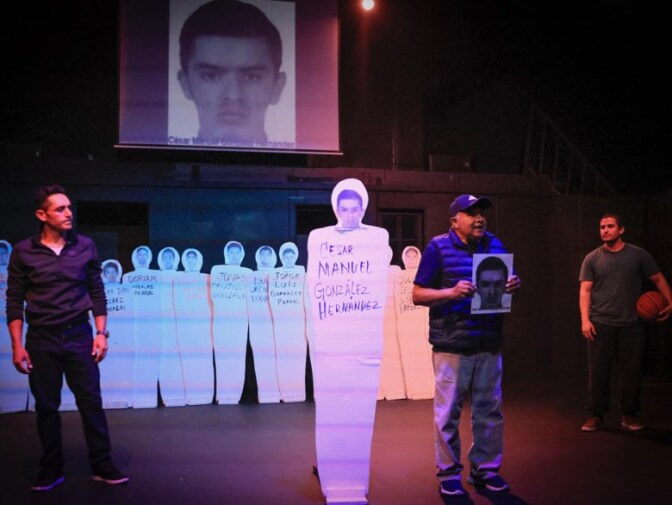 A man holds a photo on stage next to paper cutouts of bodies with photos people's faces on the heads and names written on the body.