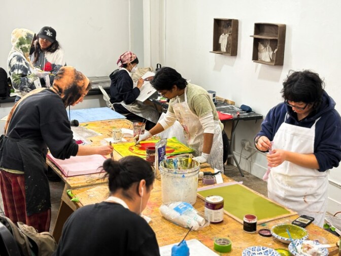 A classroom with multiple people wearing aprons painting on canvases on a table.