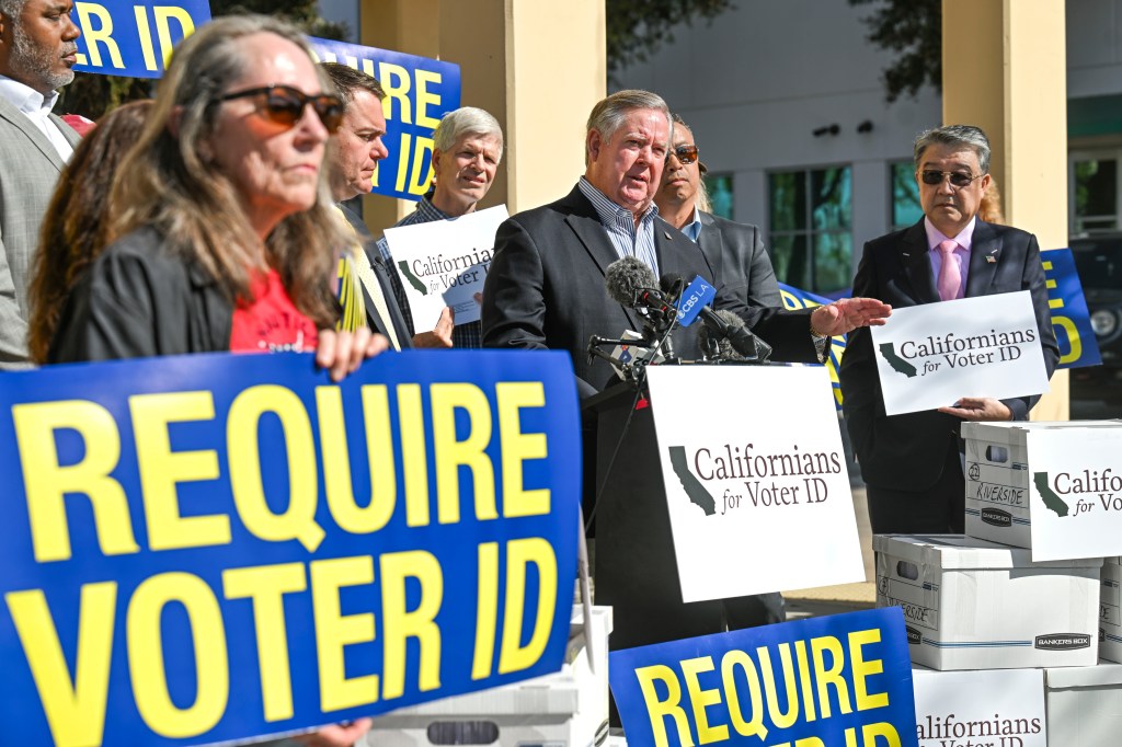 Rep. Ken Calvert speaks at a press conference announcing support for a voter ID measure, surrounded by people holding signs that say "REQUIRE VOTER ID" and "Californians for Voter ID."