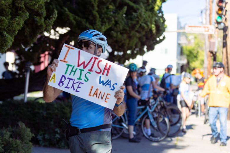 A person wearing a bike helmet holds a sign that reads "I wish this was a bike lane"