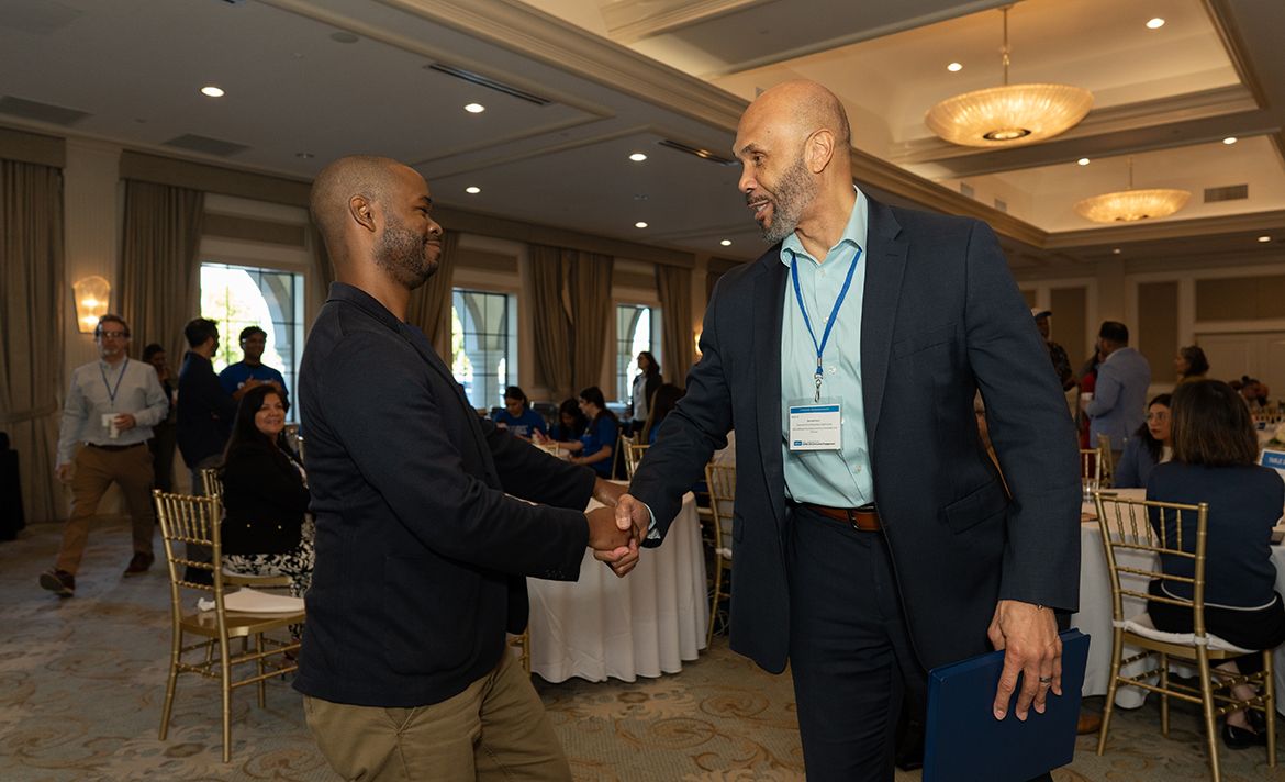 EVCP Darnell Hunt (right) speaking with panelist Corey Mathews of Liberty Hill. Two men shake hands and smile in a well-lit banquet hall during a professional event. The man on the right wears a navy blazer, light blue dress shirt, and a conference lanyard with a name badge.
