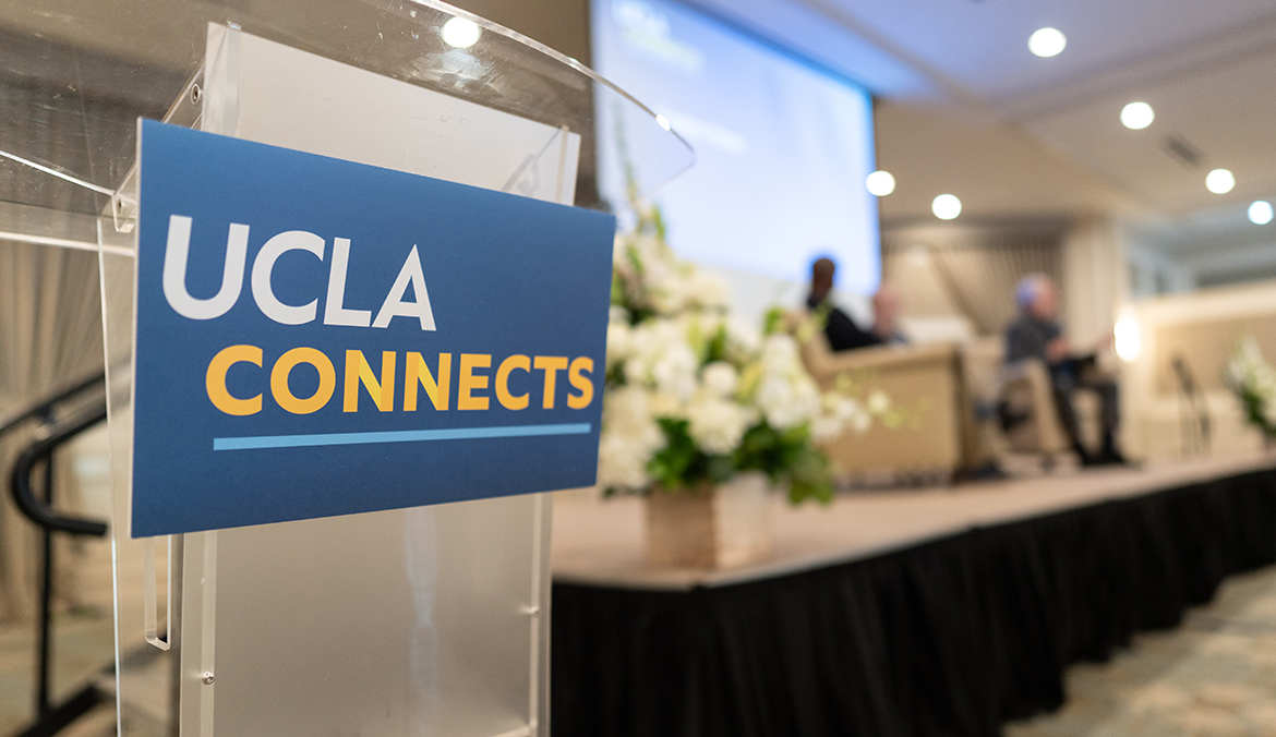 The summit exemplified UCLA Connects, a strategic initiative led by Chancellor Julio Frenk to build deeper engagement within and beyond campus. Alt text / caption: A clear acrylic podium in the foreground displays a blue sign reading "UCLA Connects" in white and gold text.