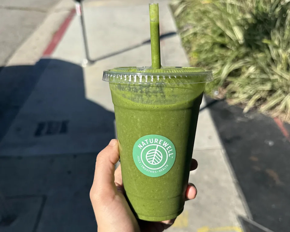 A hand holding a green smoothie with a Naturewell sticker on the front.