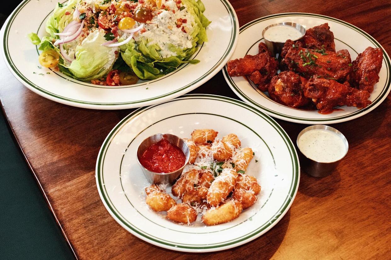 A large wedge salad, a plate of chicken wings and a bowl of fried cheese curds on a wood table