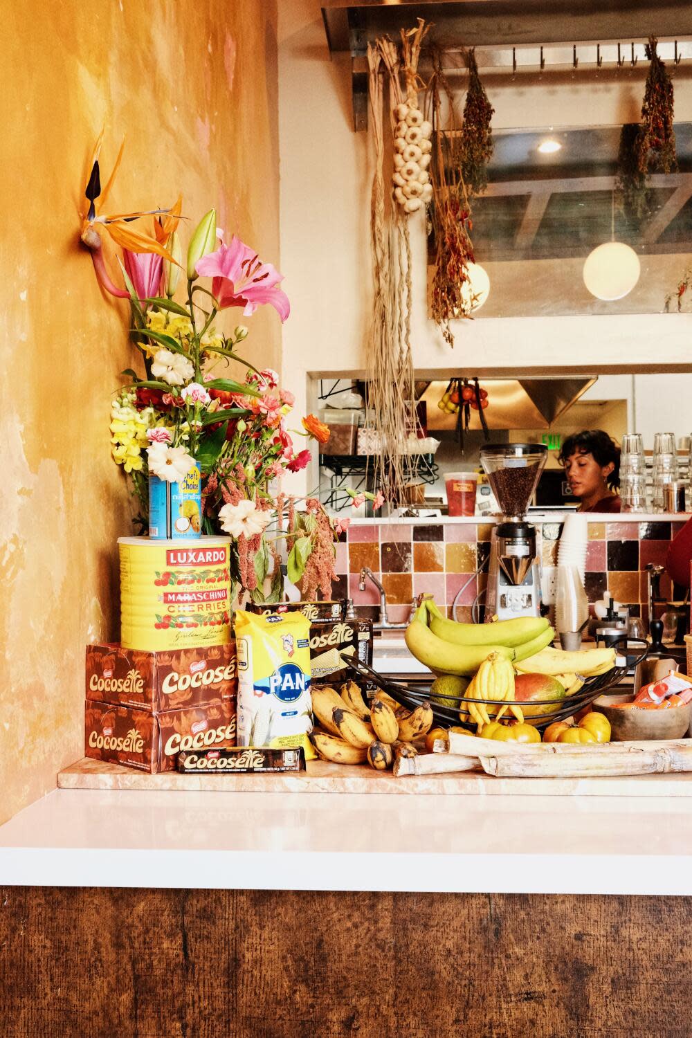 A photo of a countertop decorated with fresh flowers, bushels of bananas and imported tins. Behind, a staff member works