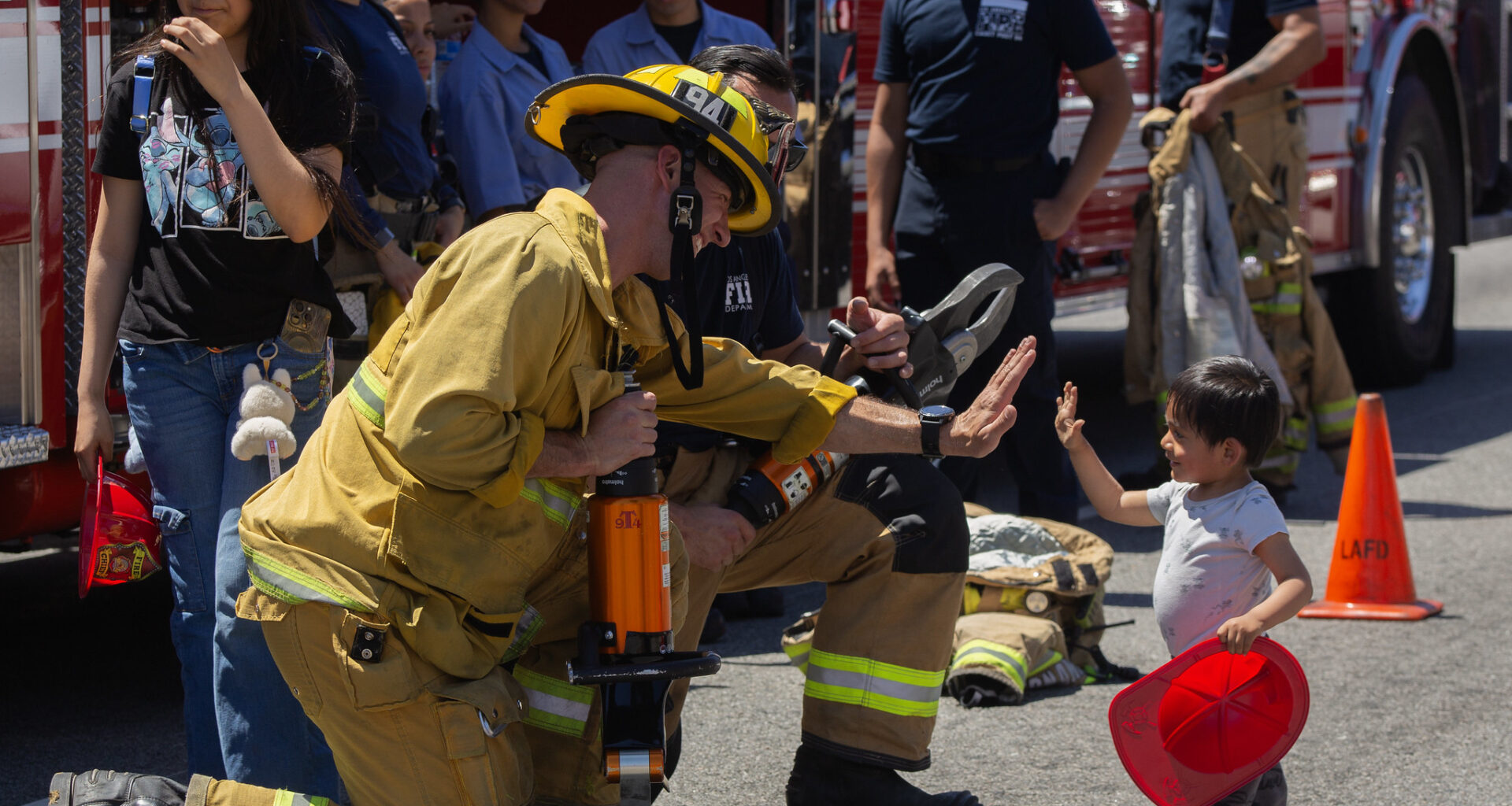 A firefighter in gear kneels, high-fiving a small child holding a red helmet. Onlookers smile. Fire truck and equipment are in the background. Festive and engaging.