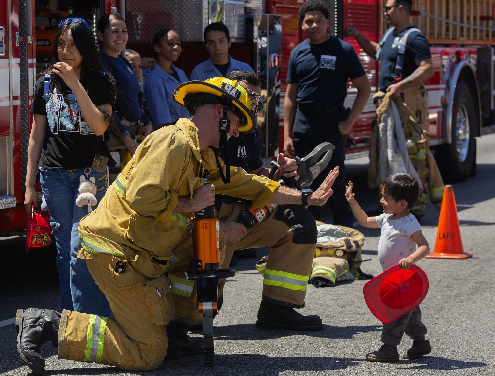 A firefighter in gear kneels, high-fiving a small child holding a red helmet. Onlookers smile. Fire truck and equipment are in the background. Festive and engaging.
