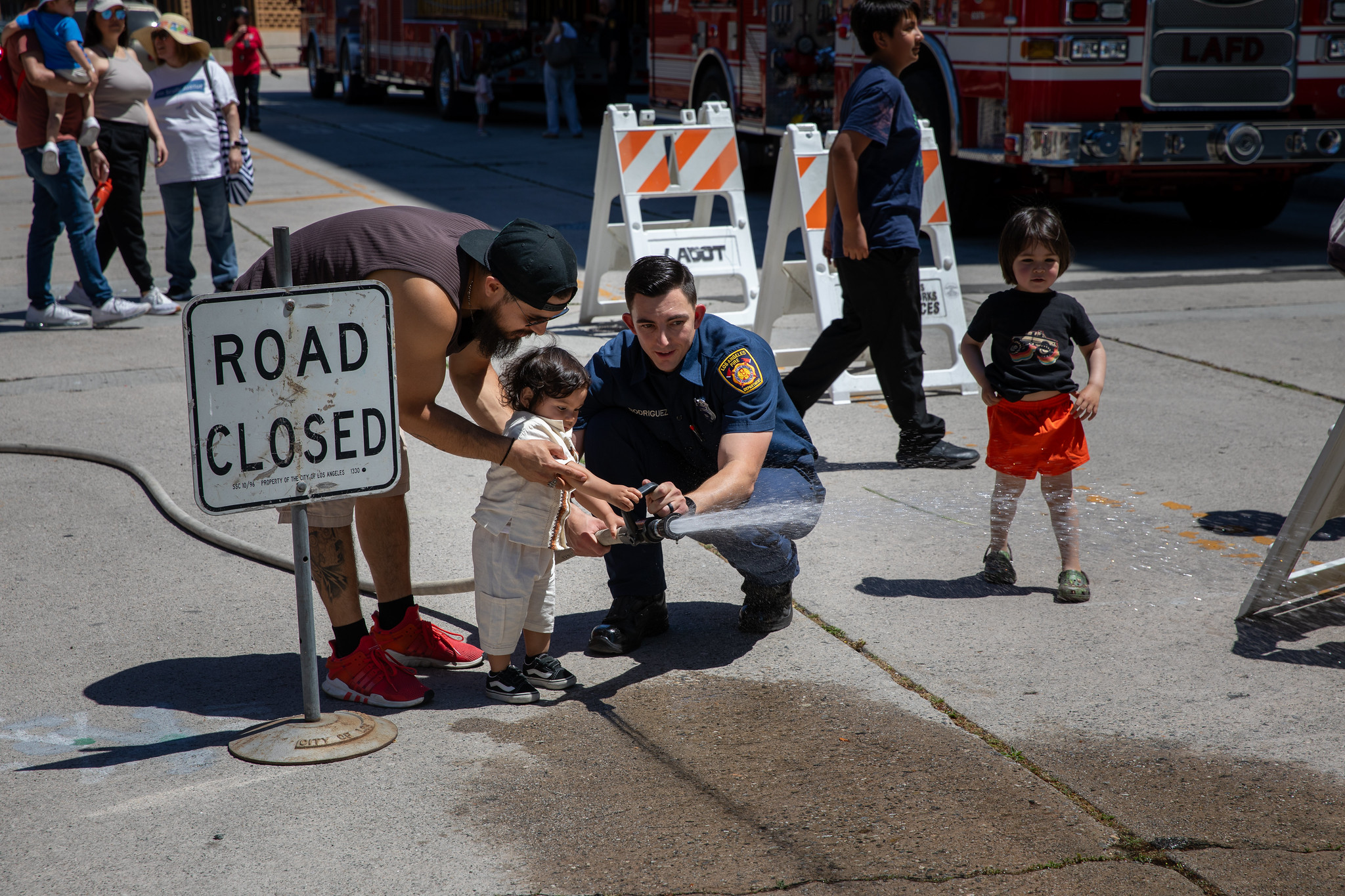 A firefighter helps a small child spray water from a hose on a sunny day. A road closed sign, fire trucks, and people are visible in the background.