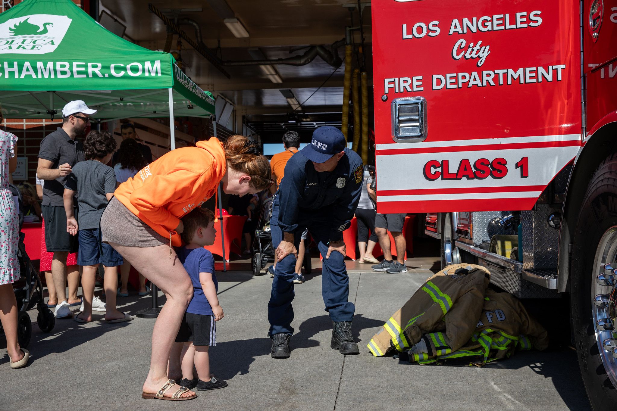 A firefighter bends down to chat with a small child and a woman in front of a red Los Angeles City fire truck. Nearby, people mingle under a green tent.