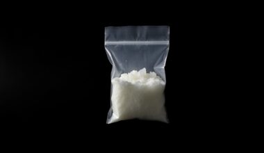 An extreme close-up photograph of a small clear plastic bag containing a white crystalline substance, lit by a harsh, direct camera flash against a pitch-black background, conceptually illustrating the discovery of suspected drugs during a probation search.