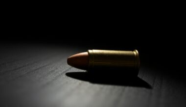 An extreme close-up photograph of a single spent bullet casing on a dark, shadowy surface, lit by a harsh, direct camera flash, conceptually representing the aftermath of a deadly shooting incident.