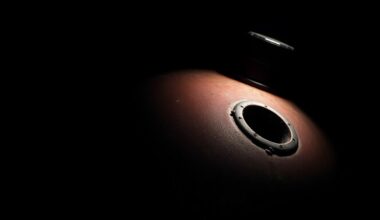 An extreme close-up photograph of a drilled gas tank, the circular hole and surrounding gasoline stain captured in stark detail against a pitch-black background, conceptually illustrating the nefarious methods used by thieves to steal fuel from vehicles.