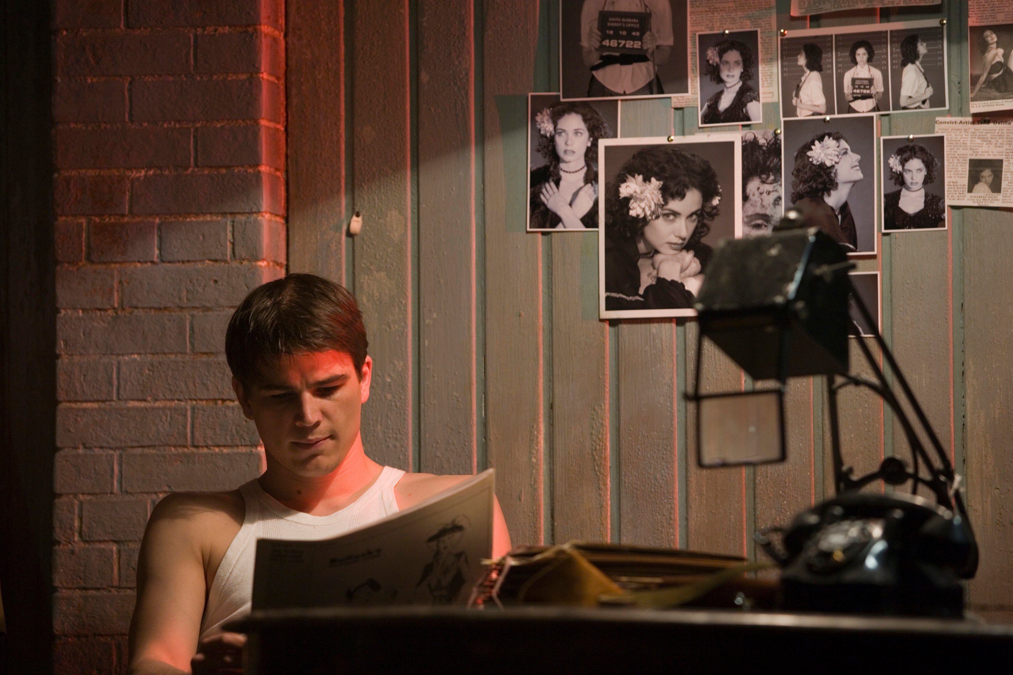 A man wearing a white tank top sits at a desk, reading a newspaper, with a wall of black-and-white glamour shots, as well as a few mug shots, behind him.