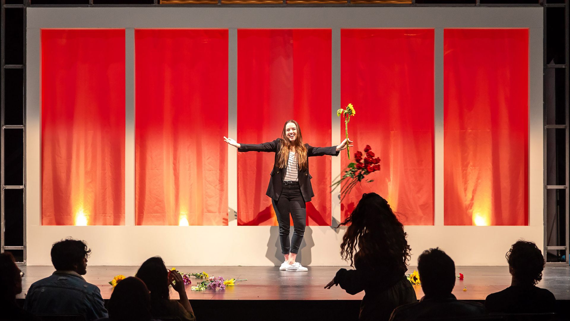 Performer holding flowers stands center stage smiling as audience members applaud.
