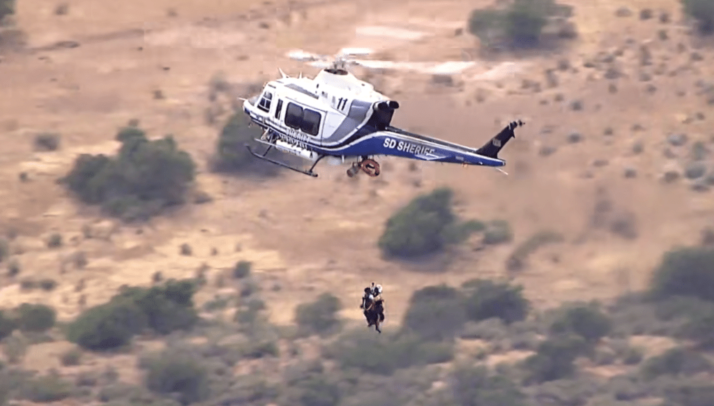 A rescue helicopter hovers above a mountainous landscape, airlifting three individuals suspended below it.