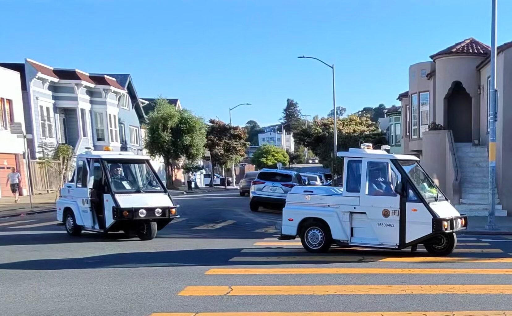 Two white parking enforcement vehicles drive through a residential intersection with houses, trees, and a yellow crosswalk visible.