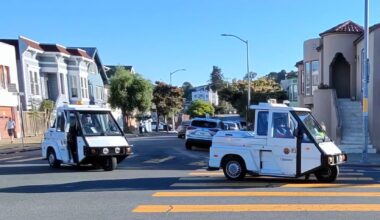 Two white parking enforcement vehicles drive through a residential intersection with houses, trees, and a yellow crosswalk visible.