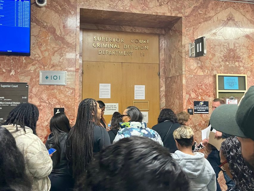 A group of people stand in a hallway outside the Superior Court Criminal Division Department 101, waiting near a closed wooden door with posted notices.