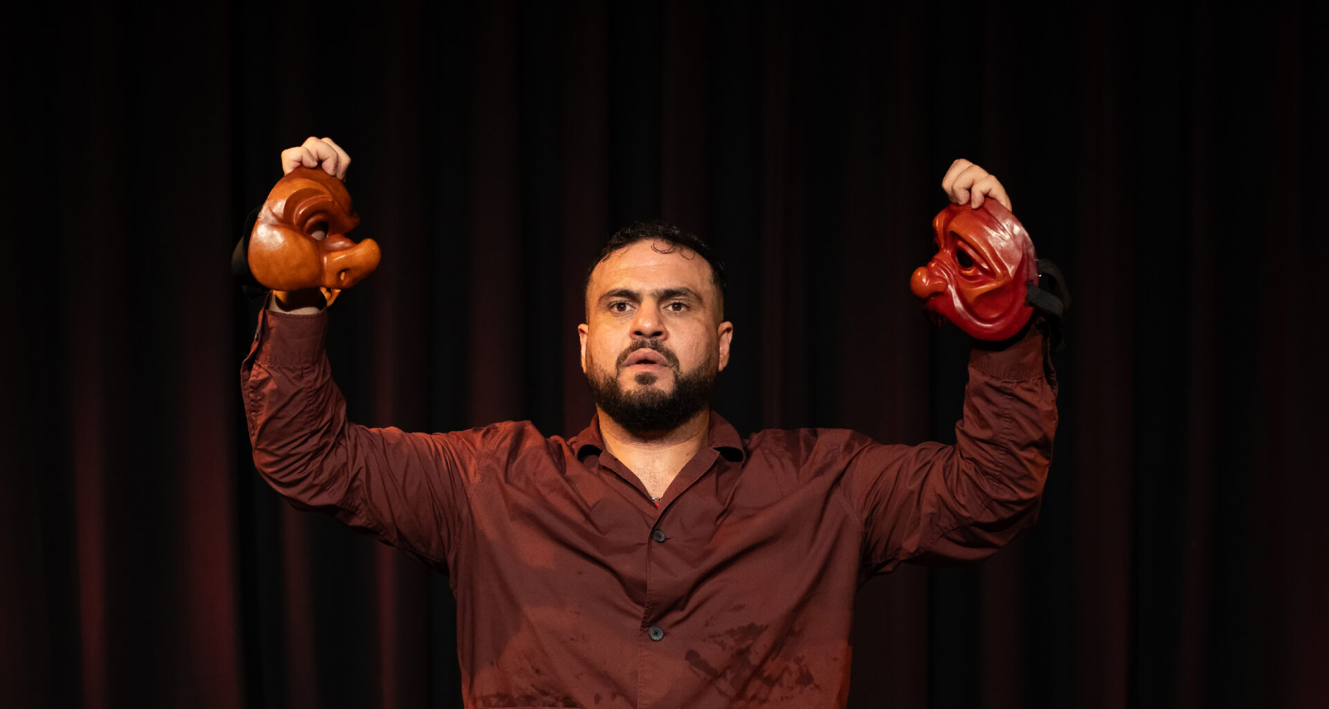 man holds two halves of mask on dark stage