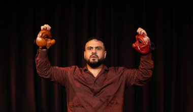 man holds two halves of mask on dark stage