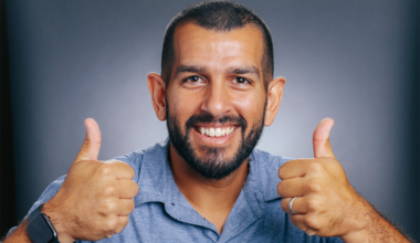 A bearded man in a blue polo shirt grins widely with two thumbs up.