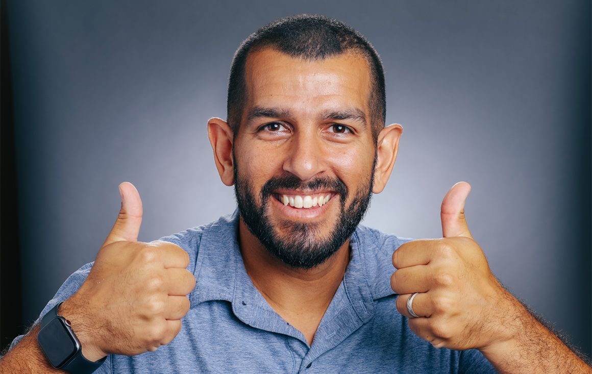 A bearded man in a blue polo shirt grins widely with two thumbs up.