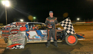 Race car driver in orange/black suit stands beside a dirt-track race car at night, holding a checkered flag; banner reads 'Feature Winner' nearby.