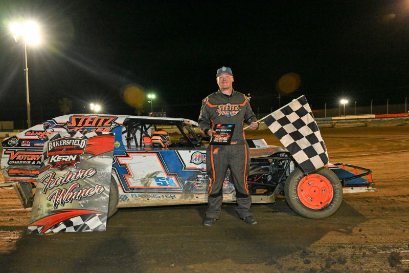 Race car driver in orange/black suit stands beside a dirt-track race car at night, holding a checkered flag; banner reads 'Feature Winner' nearby.