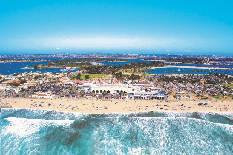 A beachside amusement park as seen from an aerial view with the ocean and sand in front of it and a broad expanse of sky behind.