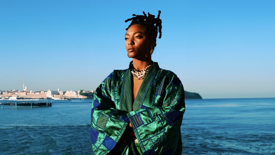 A woman wearing a green patterned robe stands with arms crossed in front of a calm blue sea and distant shoreline under a clear sky.