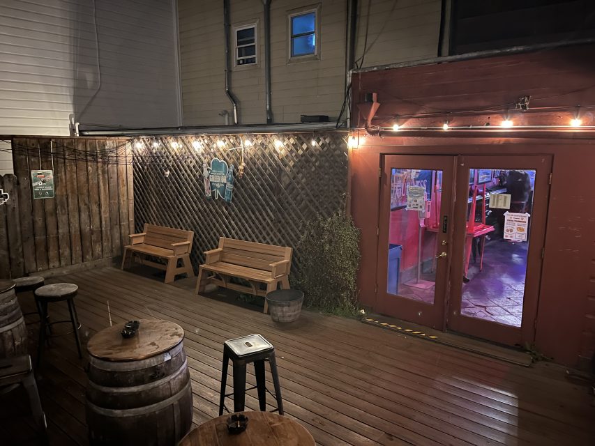 Outdoor patio at night with wooden benches, barrel tables, a stool, string lights overhead, and double doors leading into a lit indoor space.